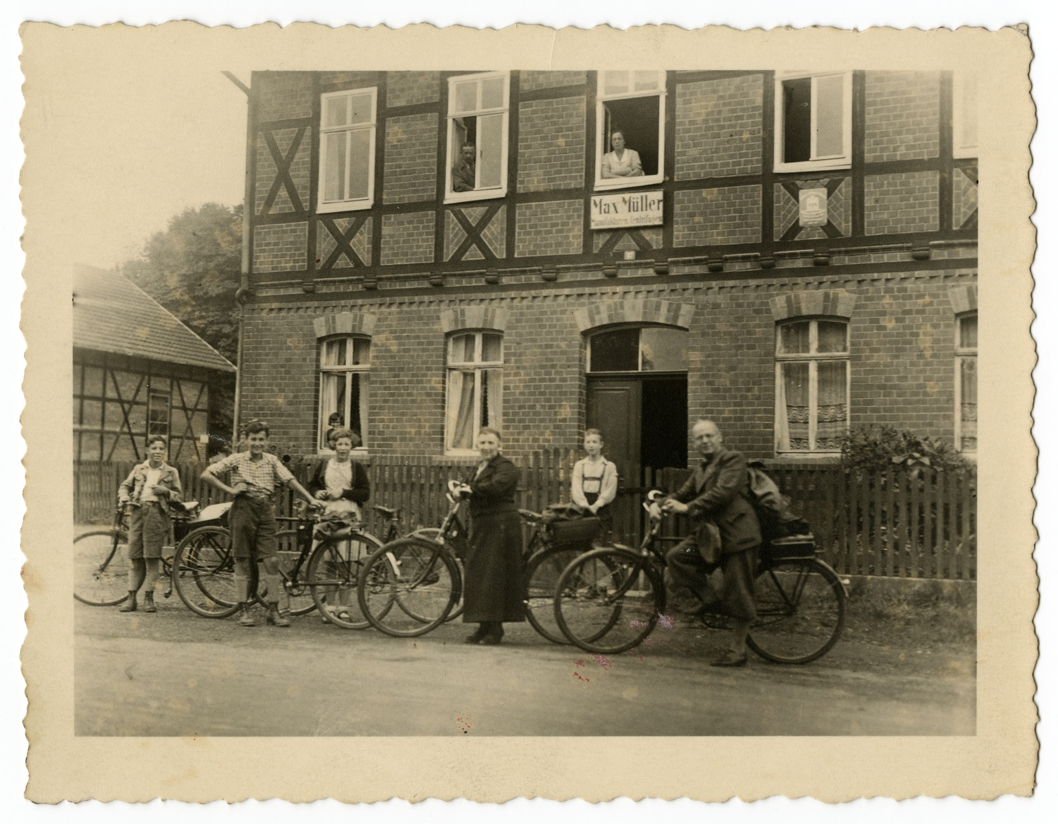 The Mueller family goes for a bicycle ride in front of the home of Sebald Mueller’s brother, Max Mueller.

Standing with their bikes from left to right are:  Norbert Mueller, his cousin Ludwig Stahl, Suse Mueller, Laura Mueller and Sebald Mueller.  The young boy in the lederhosen standing in front of the gate is Norbert’s cousin Willy Mueller, the son of Max Mueller, who appears in the second window from the left on the top floor. Max’s wife is in the next window over.