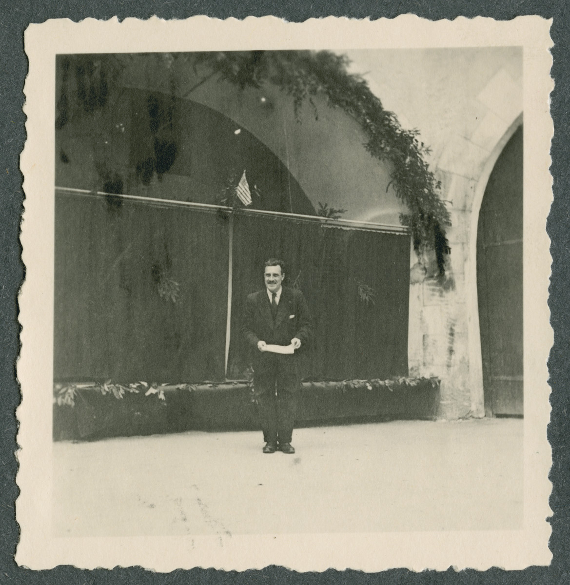 An unidentified prisoner stands underneath an American flag near the entrance to Tittmoning castle in preparation for one of the shows put on by prisoners.  Improvised stage curtains are behind him