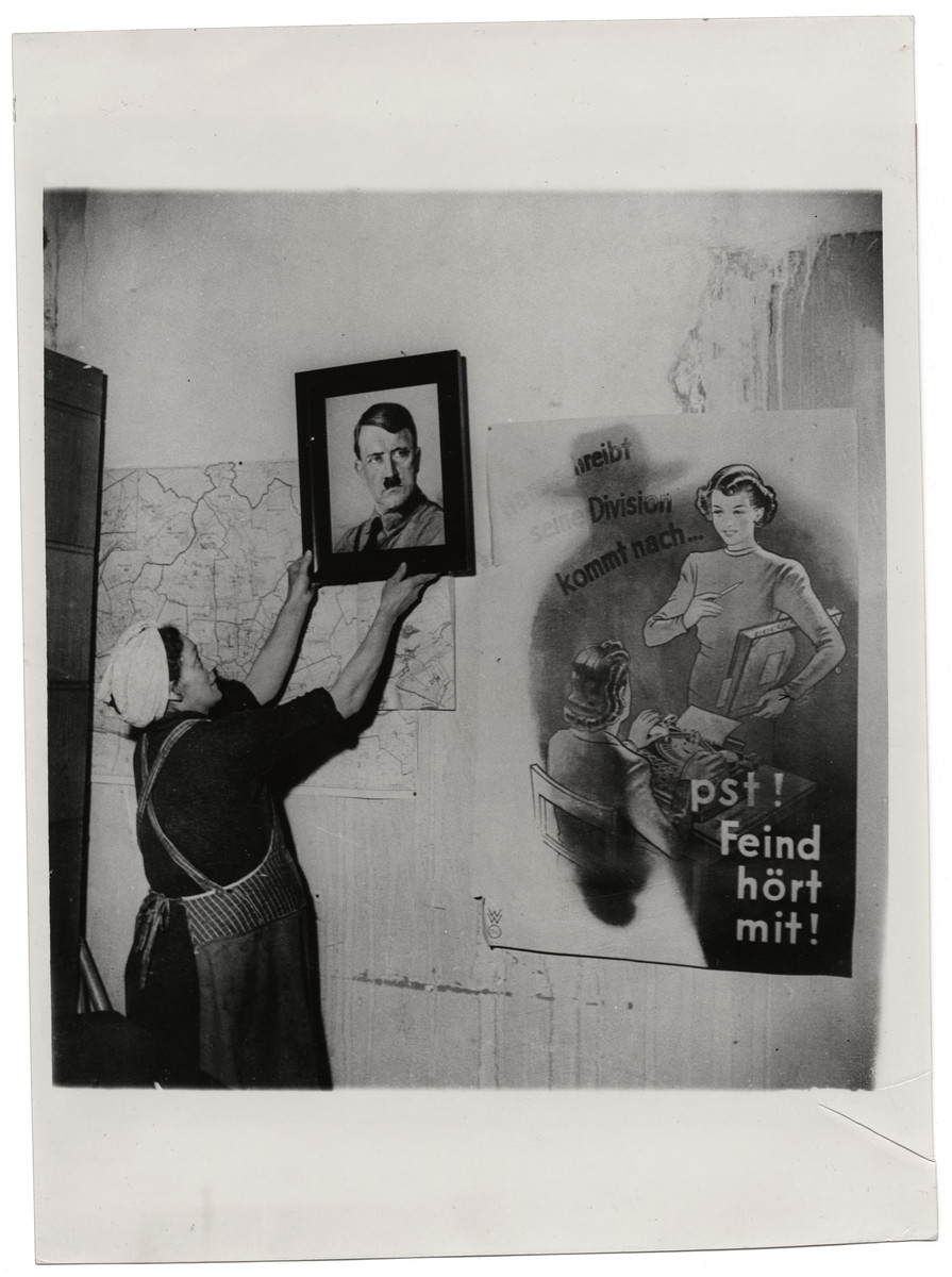 A volunteer removes a picture of Hitler in former Nazi headquarters.

Original caption reads:
Allies Take Over Frankfurt
The German population of Frankfurt- on-Main was the first large community to come under control of the U.S. Military Government. In spite of bombs and battle and repeated Nazi orders to leave, approximately 150,000 residents were still in Frankfurt when the industrial city was captured by troops of the third U. S. Army March 29,1945. 

Assisting Military Government officials are members of a citizens' council made up of members of the previous anti- Fascist parties and intellectuals of liberal views who banded together and recommended a mayor acceptable to the Allies. Frankfurt also has a new police chief appointed by the Military Government. This man was chief of the Frankfurt police until 1935, when he was ousted by the Nazis and tried on a charge of disclosing confidential information to a Jewish Social Democratic organization. His police force, which by April 2 numbered 180, has been divested of its grey- green unifroms reminiscent of the Wehrmacht. 

THIS PHOTO SHOWS: One of the women who volunteered to clean up a Protestant church, former Nazi headquarters, removes a  picture of Hitler from the wall. A propaganda poster at the right reads "Hans writes that his division is arriving at..."  "Shh, the enemy also hears". Keystone photo