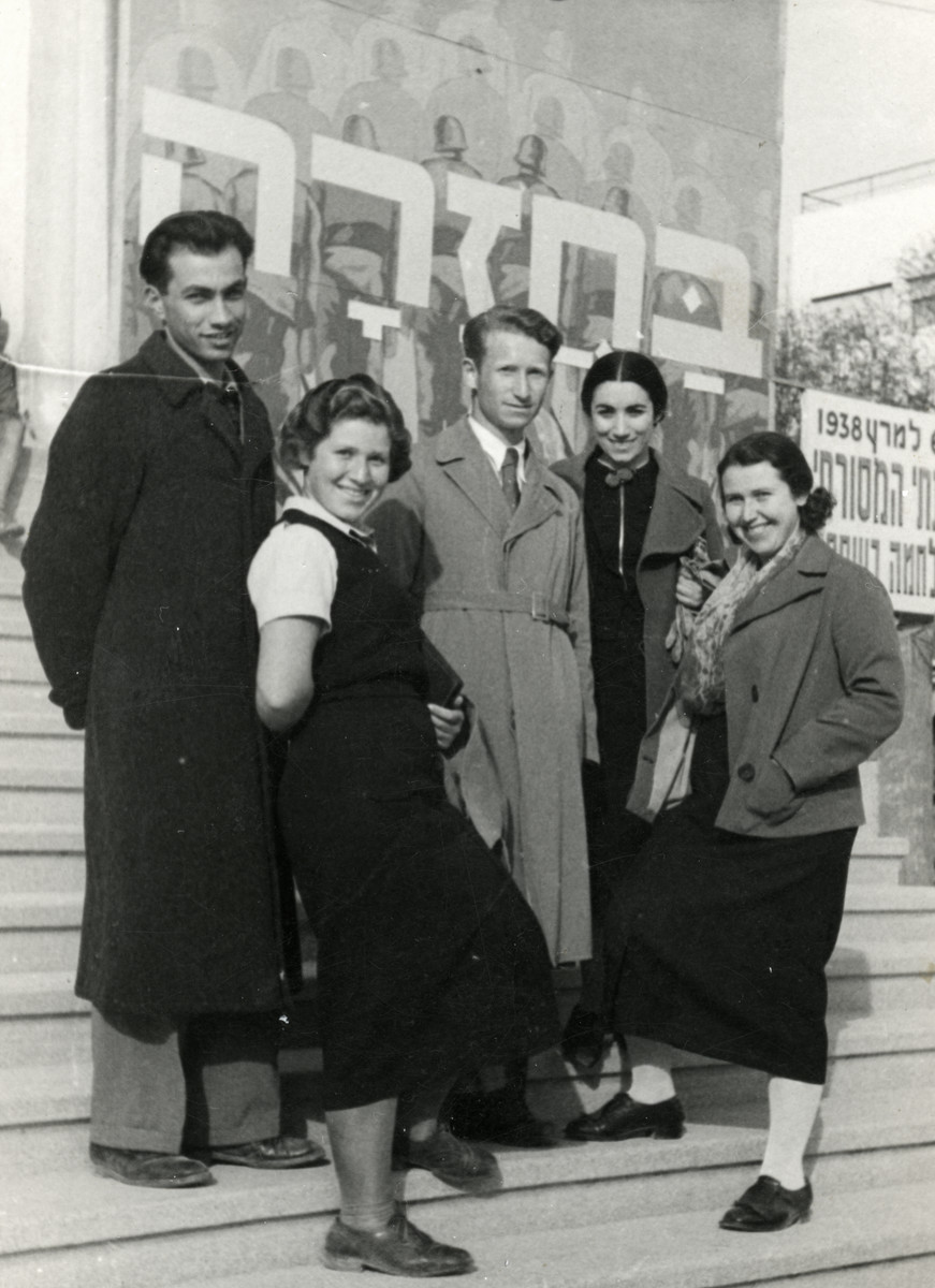 A group of young adults are pictured upon their arrival in Palestine. 

Pictured are siblings Esther (far right), Alex (middle), and Zippora Steiger (left).  The large sign behind them reads "On Return."
