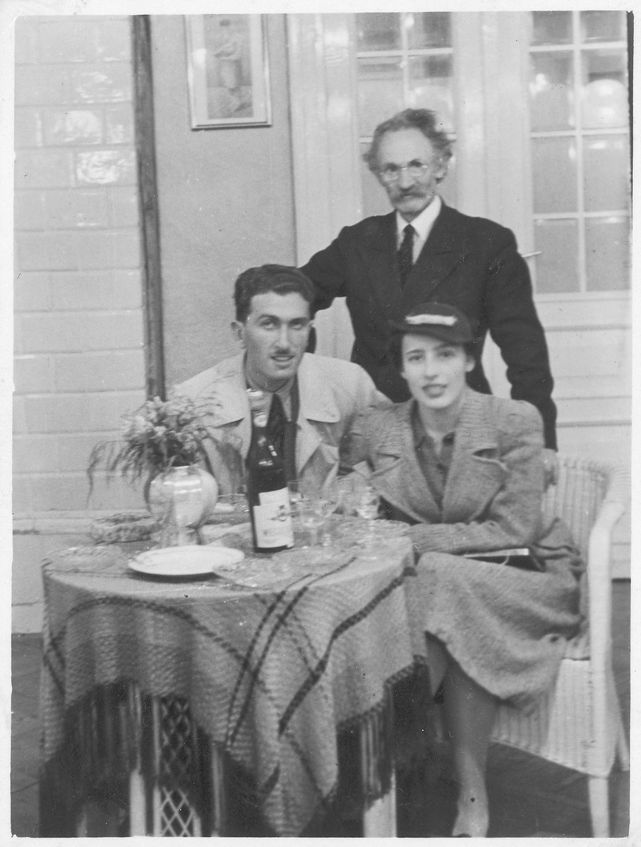 Three Polish Jews pose by a table set with wine and glasses.  

Perec Willenberg is standing.  Seated are his daughter Itta and her fiancee Shmuel Kamrat. Kamrat immigrated to Israel. Itta remained in Poland, where she perished.