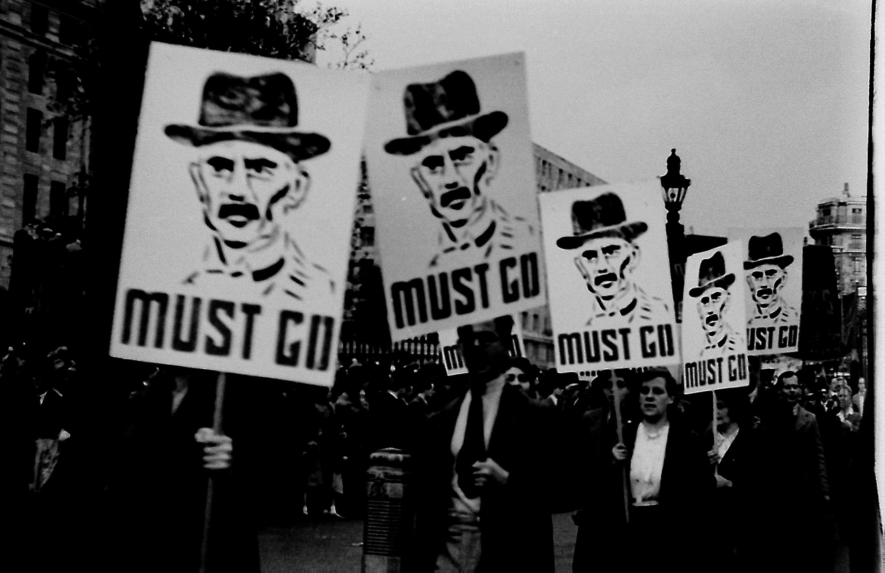 Demonstrators march through London carrying signs with a portrait of Neville Chamberlain followed by the words "Must Go" [probably right after his signing of the Munich agreement].