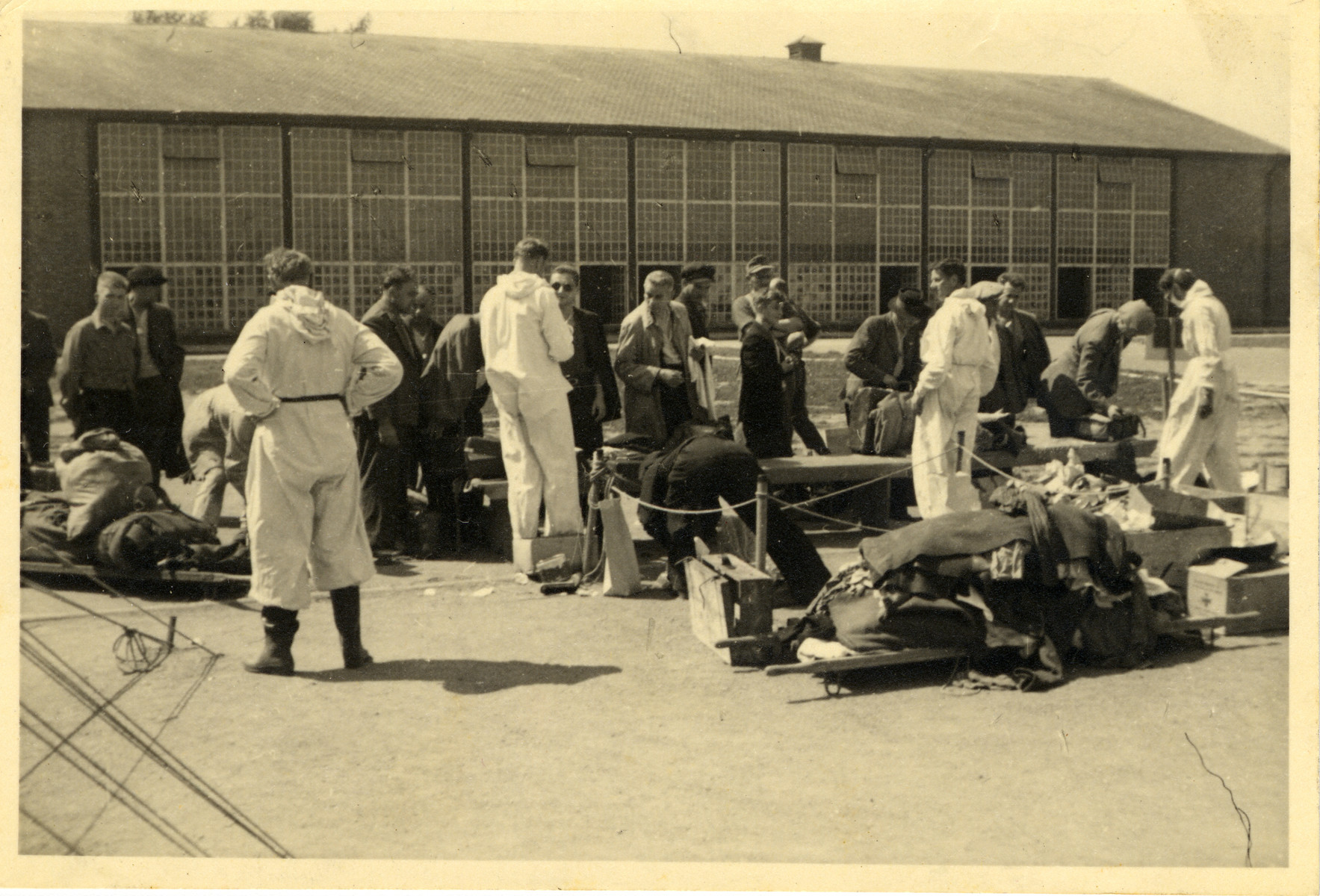 A group of men line up outside a building; they are being attended by five men dressed in white overalls.

On the floor are cardboard boxes, a suitcase, and two stretchers covered with clothes.