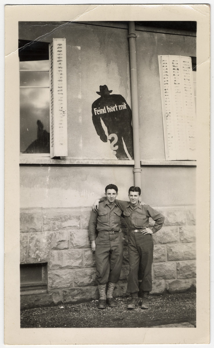 Two American soldiers pose underneath a poster that shows a lurking silhouette and a German caption that reads "The enemy is listening."