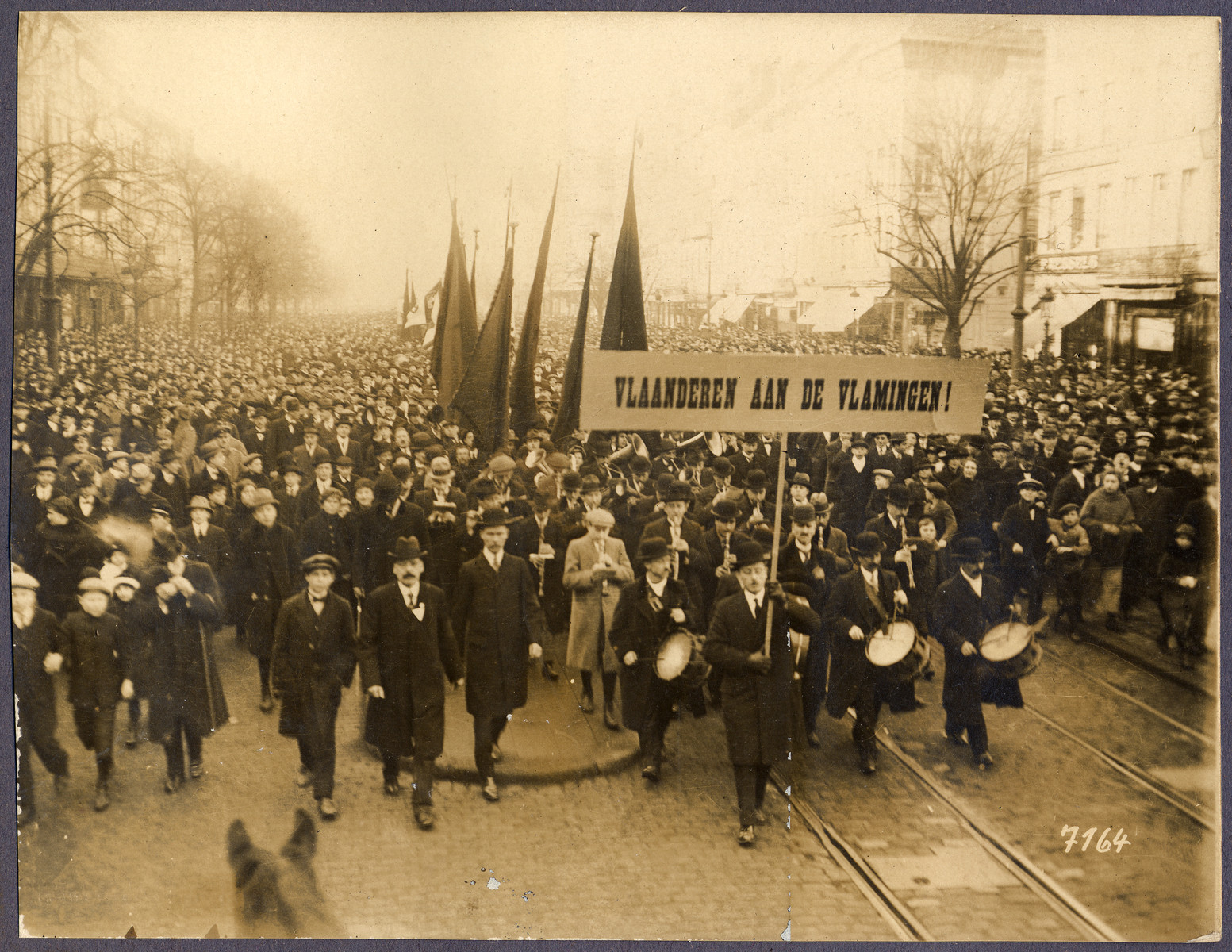 A large crowd parades through a city street carrying a sign the reads: Vlaanderen and de Vlamigen (Flanders for the Flemish).