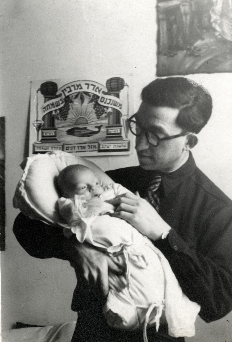 Zvi Silberman holds his newborn son in a displaced persons' camp in Austria.

Behind them is a sign that reads in Hebrew, "when Adar [the Hebrew month of Purim] comes happiness is multiplied."