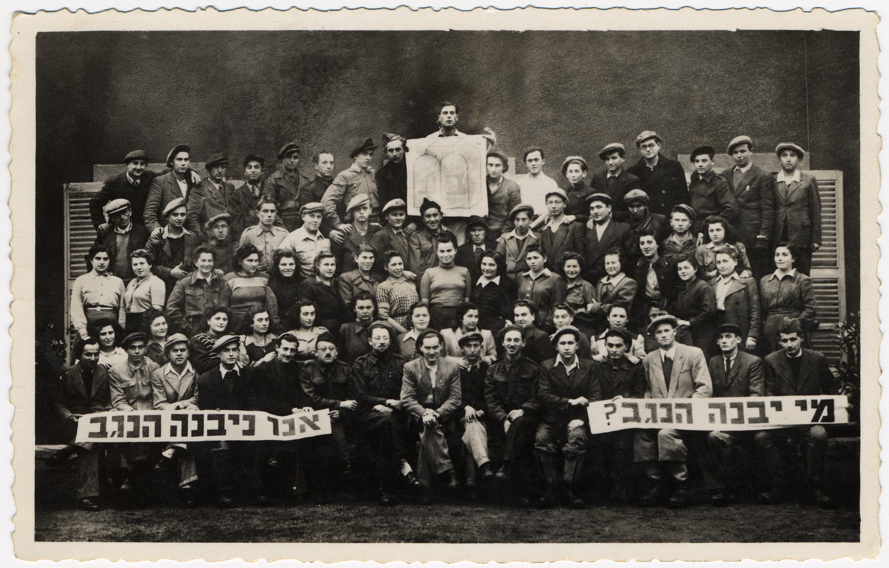 Group portrait of members of a Poal Mizrachi hachshara.

They are holding a sign with the Bnai Akiva logo and Hebrew banners which read "Who will build the Negev?  We will build the Negev".