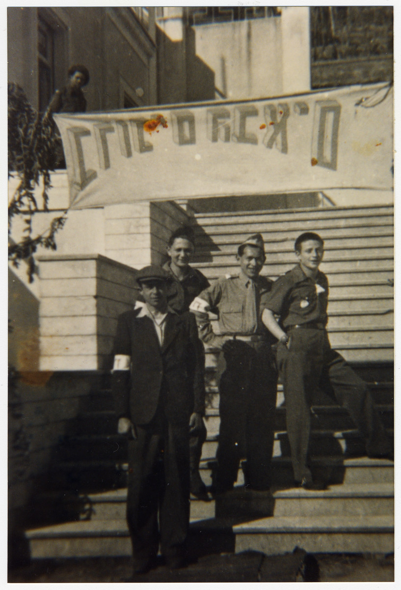 Four Jewish displaced persons stand on outdoor steps underneath a welcome banner in Kibbutz Mekor Baruch, a Poal Mizrachi fishing hachshara in Bacoli, Italy.

Among those pictured is Meier Stessel (far left).