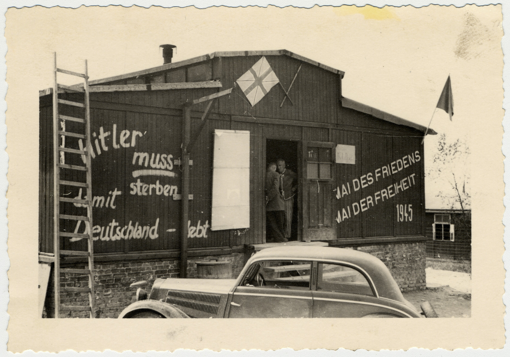 Two men stand in a doorway of the barn in Buchenwald.  

On the barn is painted, "Hitler muss sterben damit Deutschland -- lebt / Mai des Friedens/ Mai der Freiheit 1945," [Hitler must die so that Germany lives / May of Peace/ May of Freedom 1945].