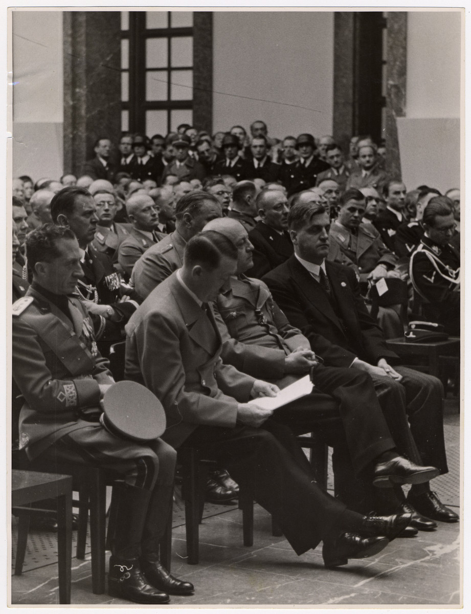 Adolf Hitler, foreign officials and other Nazi officials gather in a large room lto listen to a speaker.