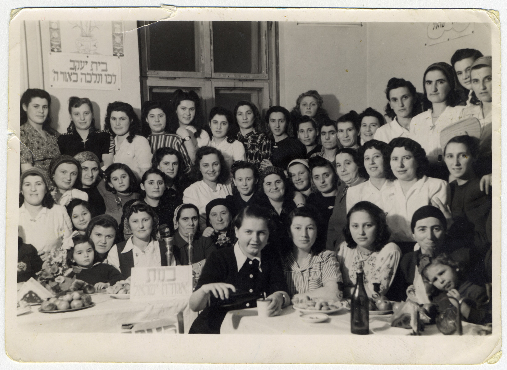 Girls from Bnot Agudat Yisrael and Beit Yaakov hold a celebration in the Bad Gastein displaced persons camp.

Pictured are Esther Ass second from the right first row.  Her sister Itka is pictured in the middle of the middle row in a light colored blouse. Also pictured is Hinde Goldberg (fifth from the right, middle row) and to her right is Yente Gelkop, and Chava Joskovitch.  Lola Schwartz nee Fried, is in the back center, with her shadow on the wall. Her sister, Magda (Matel) Gruen nee Fried,  is in the second row left, right under the woman in the printed dress.