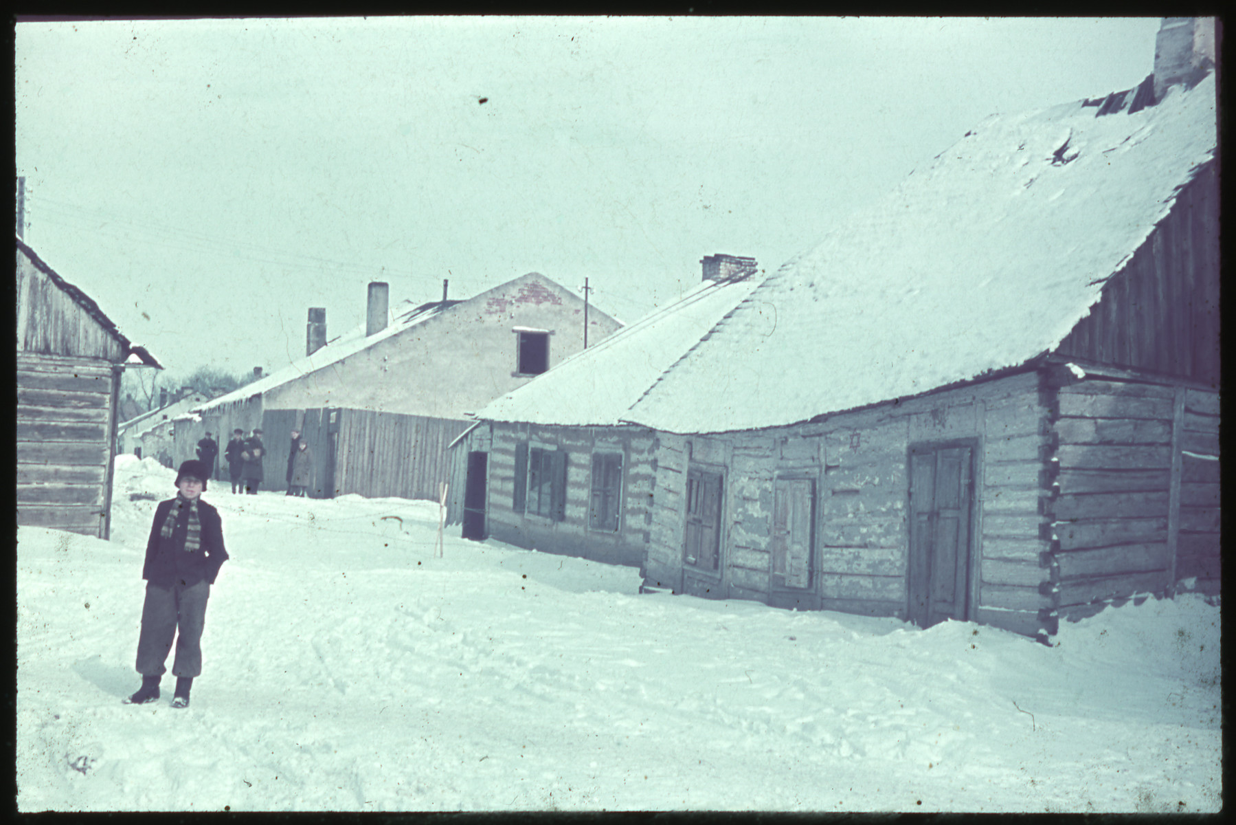 A young boy stands on a snowy road next to a building marked with a Jewish star on its exterior wall while men wearing armbands gather in the background.
