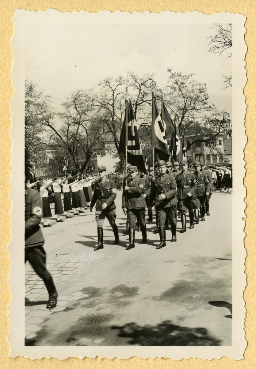 Nazis carrying flags march down a street while members of the League of German Girls stand on the sidewalk salute.