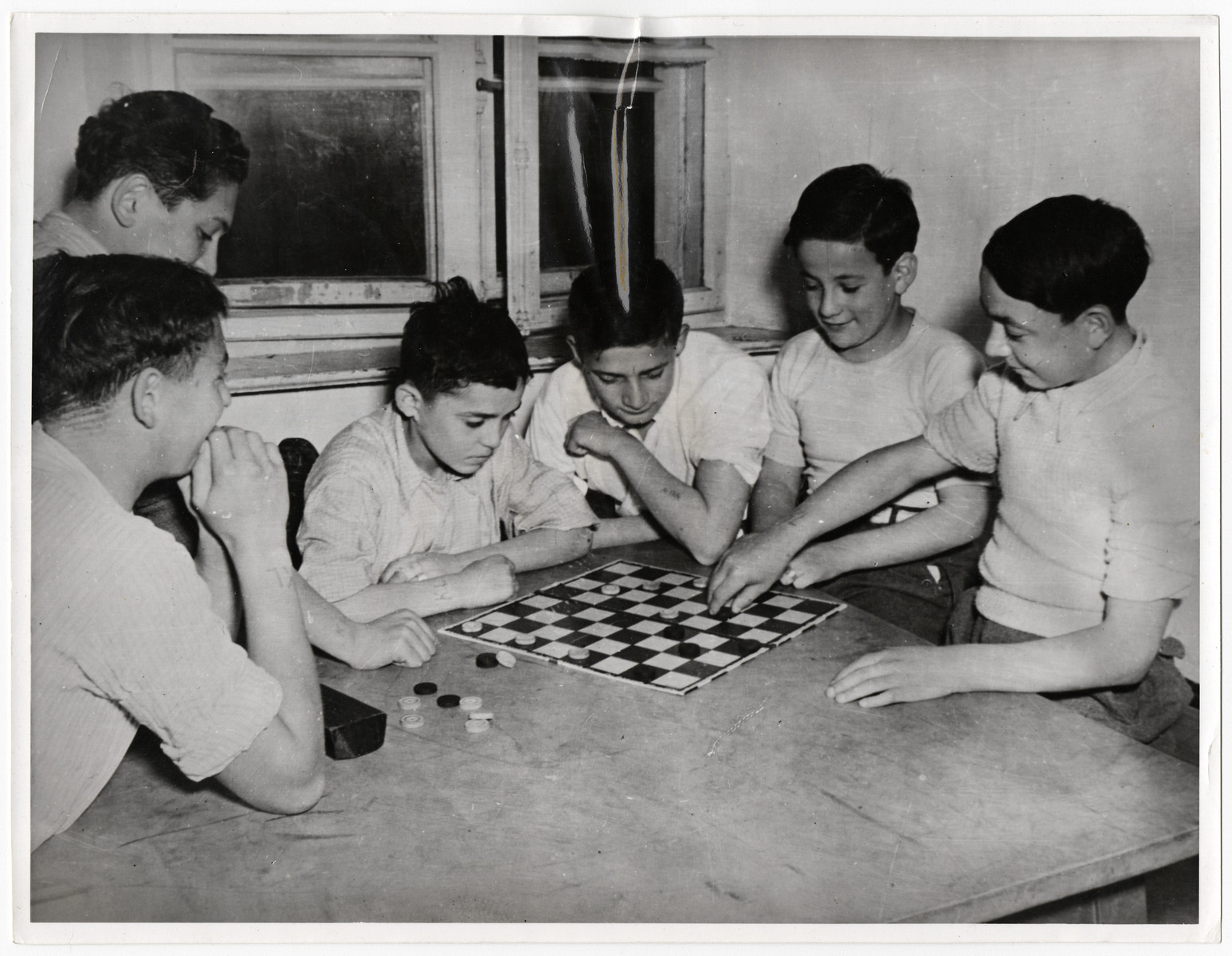 Young boys play checkers in Kloster Indersdorf children's home. Many have the initials KL tattooed on their arms.

Pictured on the left center is Sacher (later Steve) Israeler.

Original Caption: "Orphans of 12 nations have found a new home in a former nunnery at Indersdorf, Bavaria, where UNRRA (United Nations Relief  and Rehibilitation Administration) recently opened an international refuge for children whose parents were killed or lost during the war were sent from Nazi foreign labor and concentration camps to this institution. Two-thirds of the group are Polish and Jewish. Some are too young to remember their parents and some do not know their own names. Most of them from concentration camps have no other identification than a number and the letters 'KL' (Konzentrations Lager) marked on their skins. When children arrive at the institution, they are deloused, bathed and given clean clothes. Medical attendants then innoculate them against typhoid fever, diptheria and smallpox. The infants are cared for by nurses while older children begin elementary education in their own language under teachers of the Allied nations."