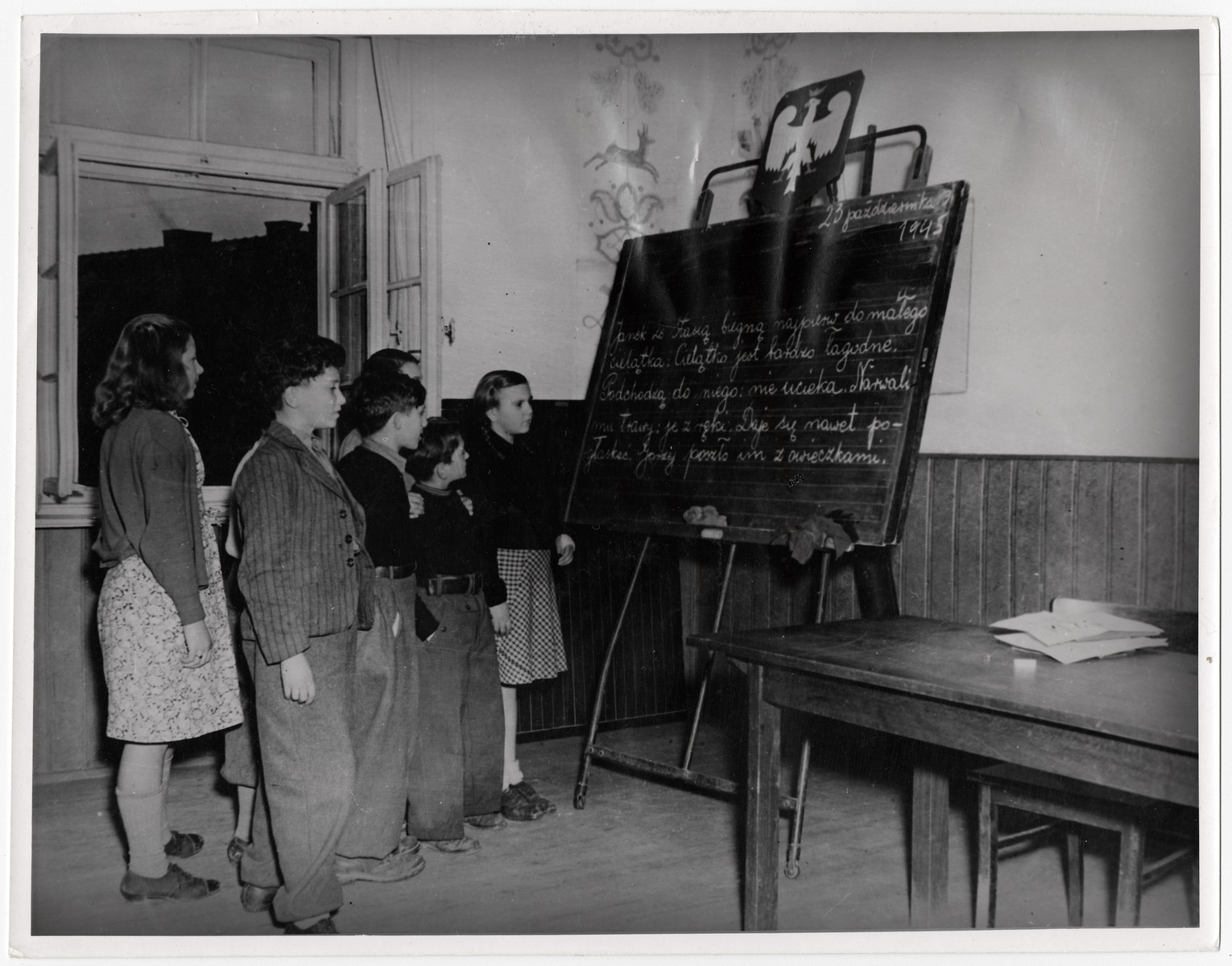 Polish children study a lesson in their native language in the Kloster Indersdorf children's home.

Original Caption: "Orphans of 12 nations have found a new home in a former nunnery at Indersdorf, Bavaria, where UNRRA (United Nations Relief  and Rehibilitation Administration) recently opened an international refuge for children whos parents were killed or lost during the war were sent from Nazi foreign labor and concentration camps to this institution. Two-thirds of the group are Polish and Jewish. Some are too young to remember their parents and some do not know their own names. Most of them from concentration camps have no other identification than a number and the letters 'KL' (Konzentrations Lager) marked on their skins. When children arrive at the institution, they are deloused, bathed and given clean cloths. Medical attendants than innoculate them against typhoid fever, diptheria and smallpox. The infants are cared for by nurses while older children begin elementary education in their own language under teeachers of the Allied nations."