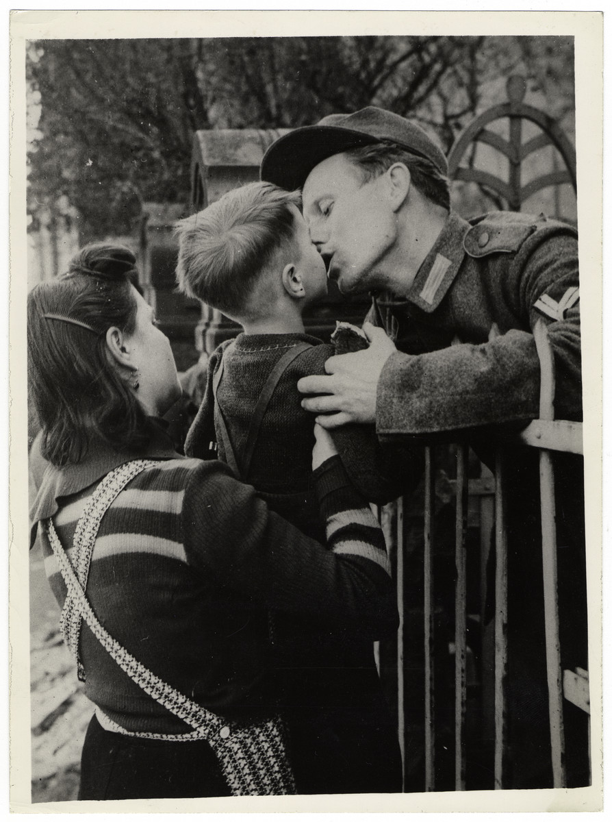 A captured German POW kisses his wife and child farewell.

Original Caption: "A German soldier, captured by the Third U.S. Army, leans over the railings of a temporary prisoner-of-war cage in Worms to bid farewell to his wife and son before being transported to the rear. Worms, nine miles north of Ludwigsshafen on the west bank of the Rhine, fell to the Americans March 20, 1945. from March 14 to25, the third U.S. Army took 76,997 prisoners."