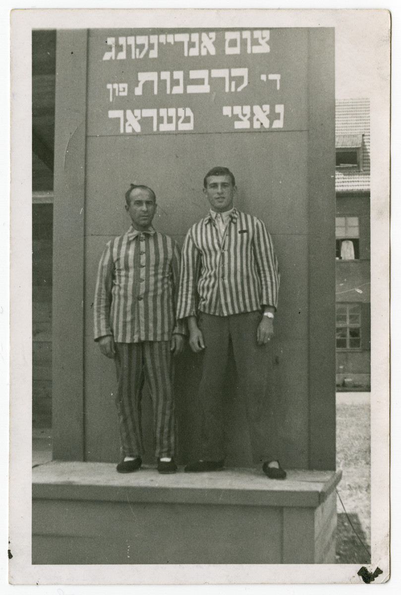 Two Jewish displaced persons wearing their concentration camp uniform pose in front of a memorial to the victims of the Holocaust.

The Yiddish memorial reads: "As a memory to those sacrificed to Nazi terror."
