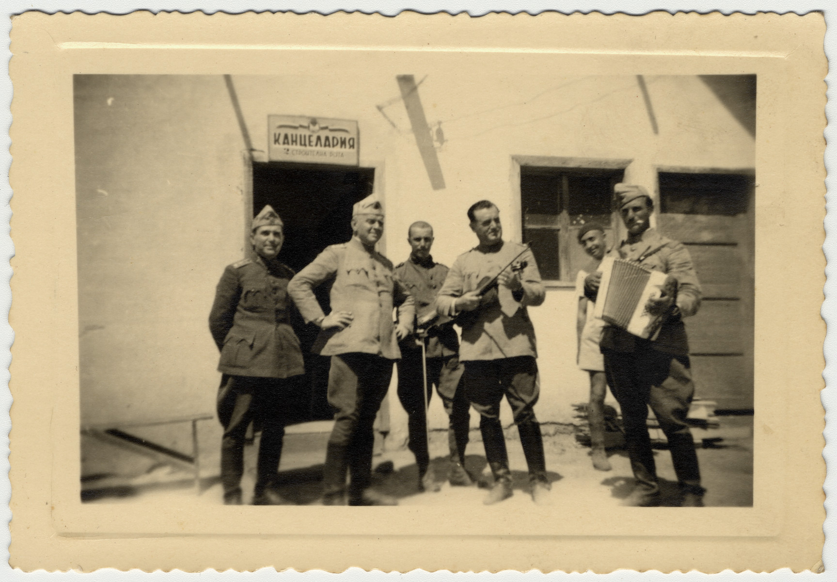 Bulgarian officers play musical instruments where they are stationed at a labor brigade in front of a building with a sign that reads "Chancellery".

Pictured in the center with a violin is Nissim Farhi.  Though Jewish, he was allowed to wear his uniform and socialize with other officers.