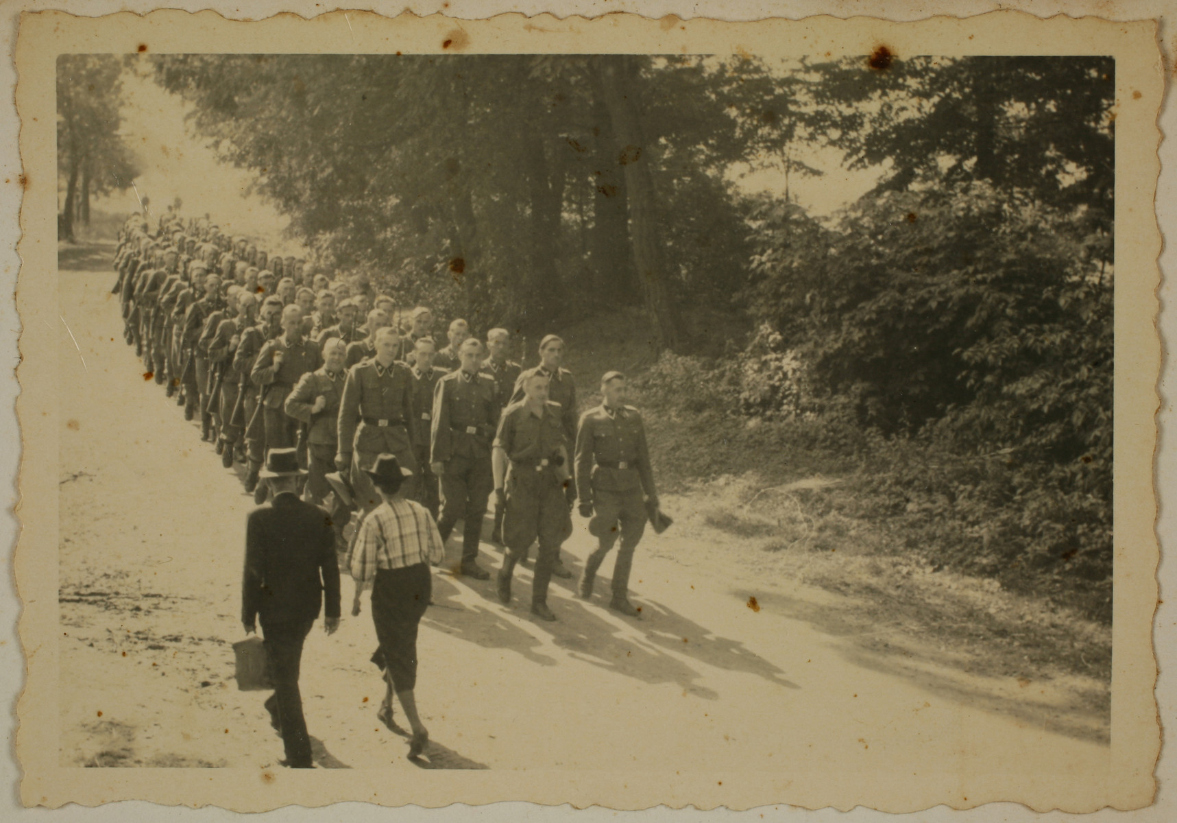SS officers and enlisted men march to shooting practice. Two civilians walk in the opposite direction.

The original caption reads "Mit den Kdtr.-Stab auf dem Marsch zum Schiessstand" (on the march with the commandant's office staff to the firing range).
