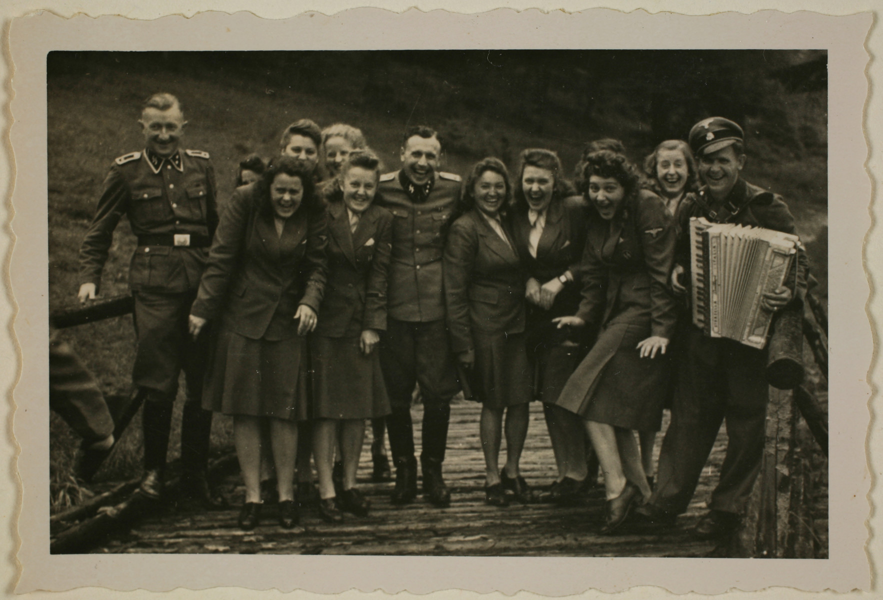 Nazi officers and female auxiliaries (Helferinnen) pose on a wooden bridge in Solahuette. The man on the right carries an accordion. 

Karl Hoecker is pictured in the center.

The original caption "Regen aus heiteren Himmel" [Suddenly, it started to rain.]