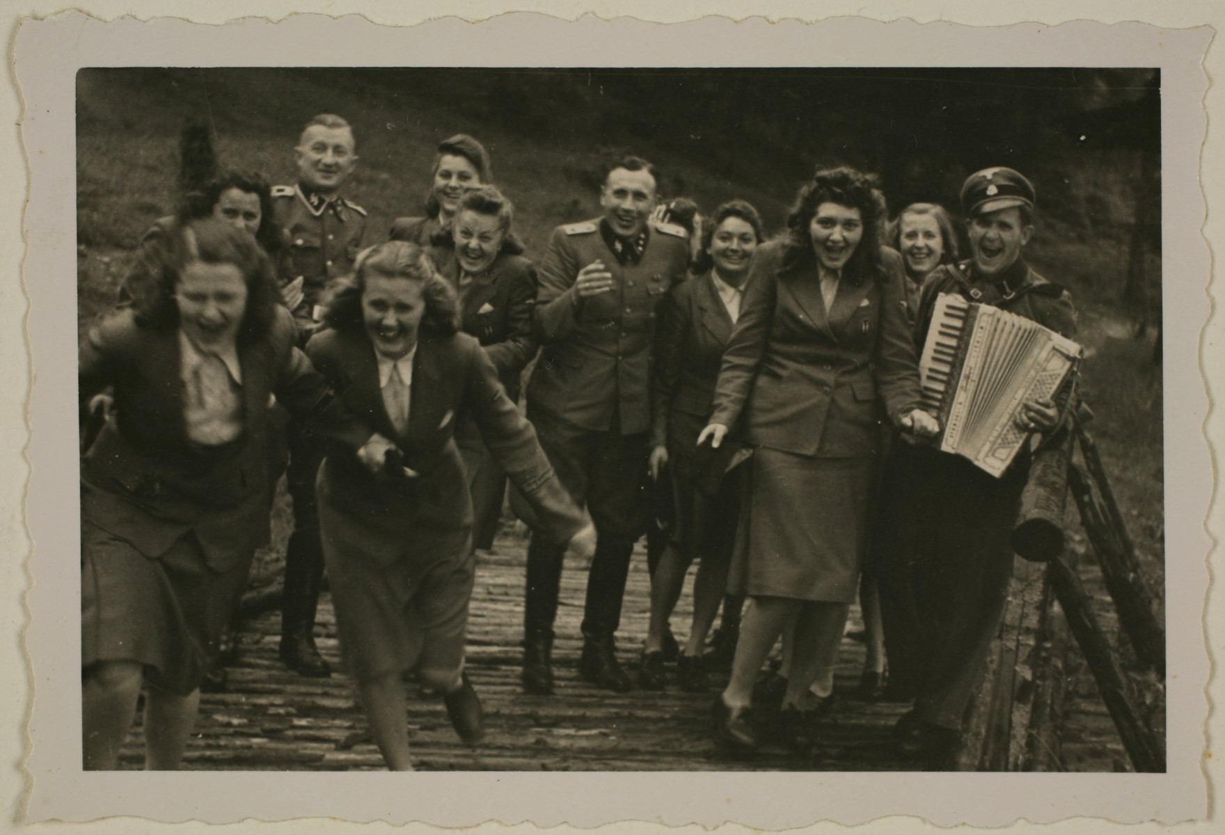 Nazi officers and female auxiliaries (Helferinnen) run down a wooden bridge in Solahuette.  The man on the right carries an accordion.

Karl Hoecker is pictured in the center.

The original caption "Regen aus heiteren Himmel" [Suddenly, it started to rain.]