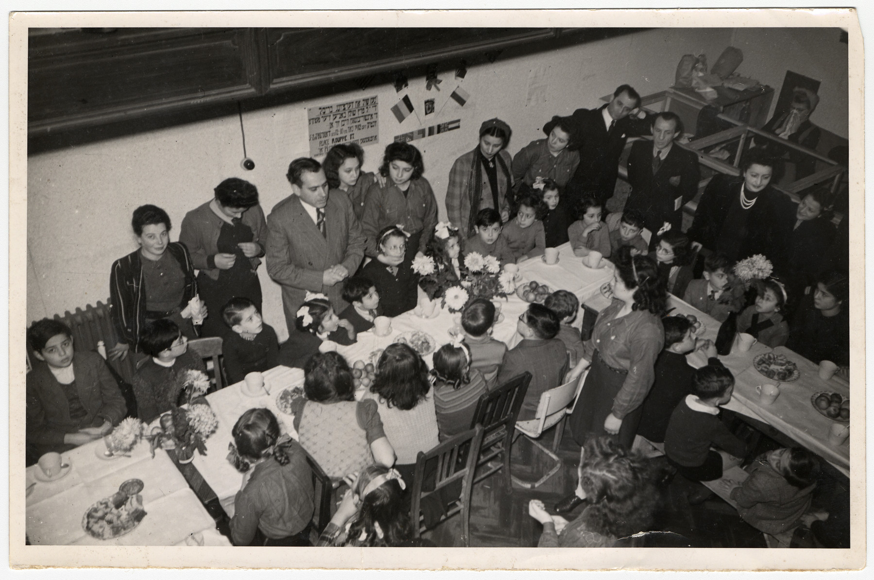 Children in a postwar children's home gather around long tables set with fruit and decorated with flowers.  Hebrew signs hang on the walls.

Among those pictured is Fela Perleman (standing second from the left).
