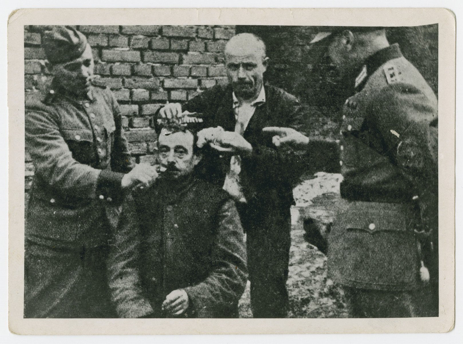 German soldiers force a man to shave the beard and hair of another [probably a religious Jew] in an act of public humiliation.