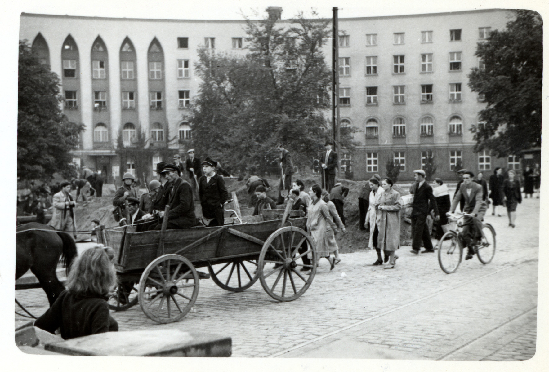 Polish soldiers and civilians dig anti-tank trenches to defend Warsaw from the oncoming German army while other civilians pass by them on the street.