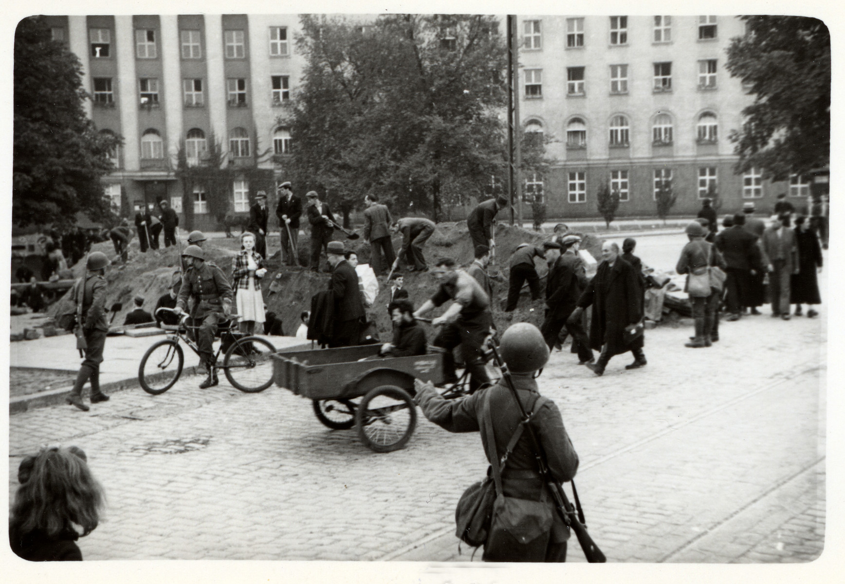 Poles dig anti-tank trenches under the supervision of the Polish army to slow down the progress of the German army,  while other civilians pass by them on the street.