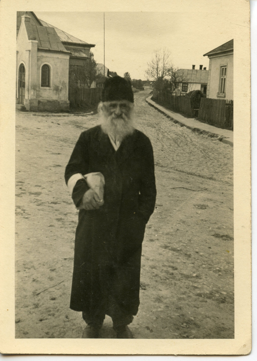 An elderly religious Jew wearing an armband carries a  bundle in an otherwise deserted road.