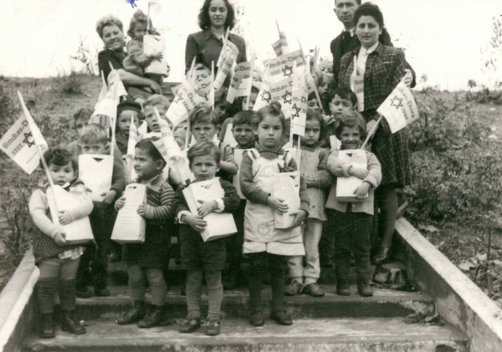 Young children carry small bags and flags during a Simchat Torah celebration in the Schlachtensee displaced persons camp.

Clara is the little girl being held by the woman on the far left, back row.