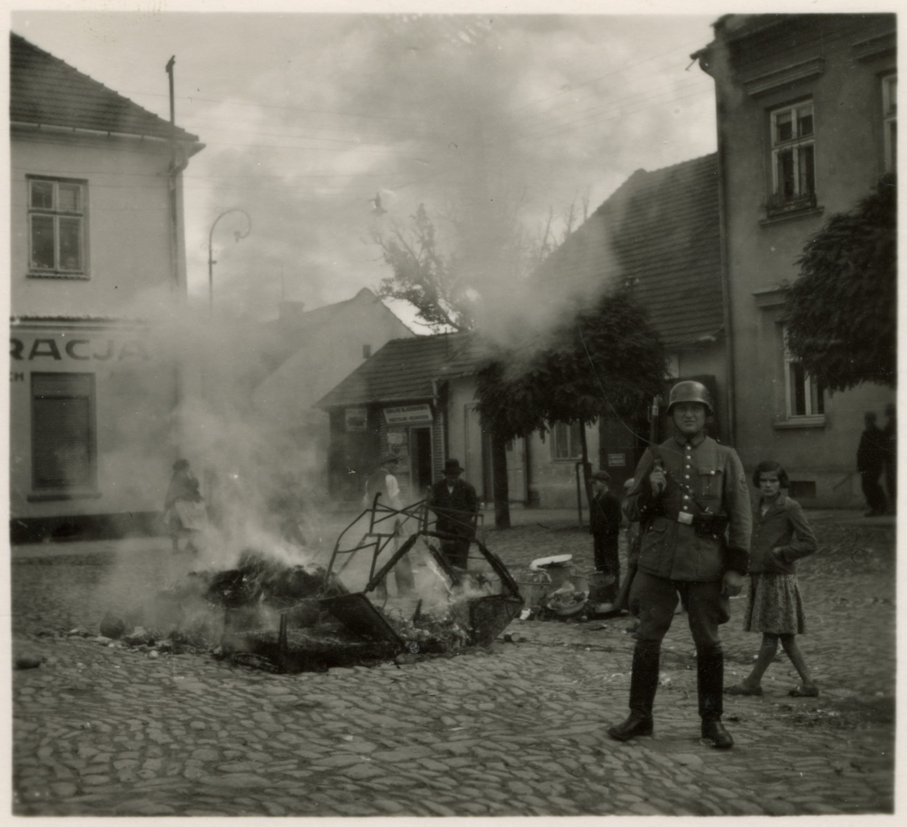 Propaganda photo showing a German soldier guarding a bonfire of burning mattresses.

The original caption reads: "Roach and lice-ridden Jewish beds being burned in the market square in Myslenice."

The photograph was part of a series of stereographic prints depicting the occupation of Poland entitled "Die Soldaten des Fuehrers im Felde."  (The Fuhrer's soldiers in the field.  The photographs were mass produced and published by Raumbild-Verlag-Otto Schönstein.