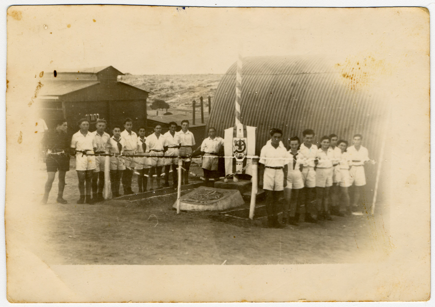 Group portrait of members of Gordonia Zionist Youth movement standing next to a flag pole in the Cyprus internment camp. 

Pictured second from the right is Bella  Bemocha (later Sajovic).