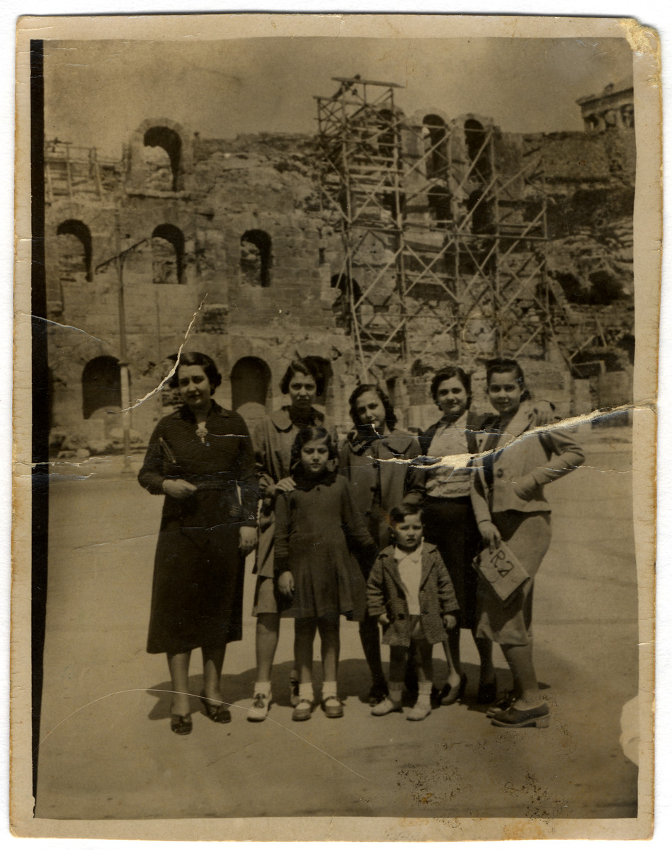 A  Greek Jewish family poses in front of a building in ruins. 

Among those pictured are Claire Jesurun (later Elhai), and her siblings Gilda, Renee, Susee, and Ralph.