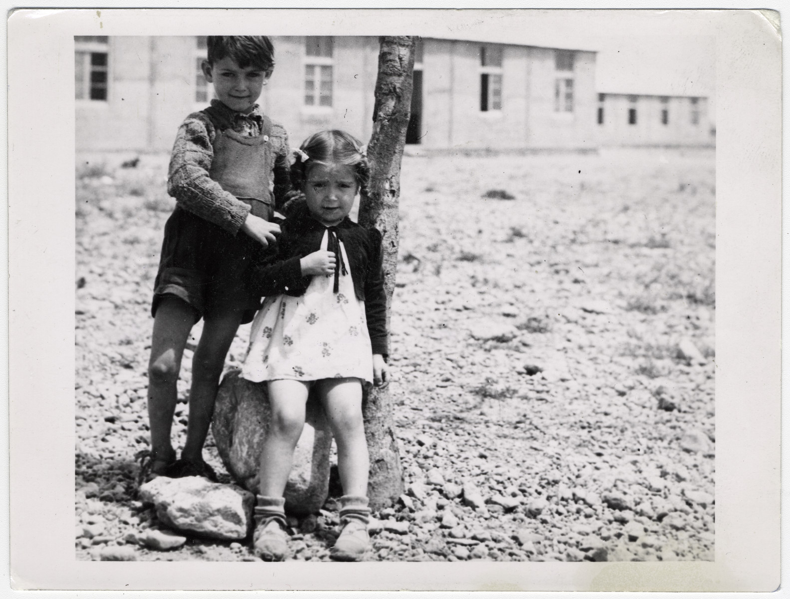 A Spanish boy and girl stand outside next to a tree, in front of a school building at Rivesaltes. 

Original caption reads: "Two Spaniards of the nursery school."