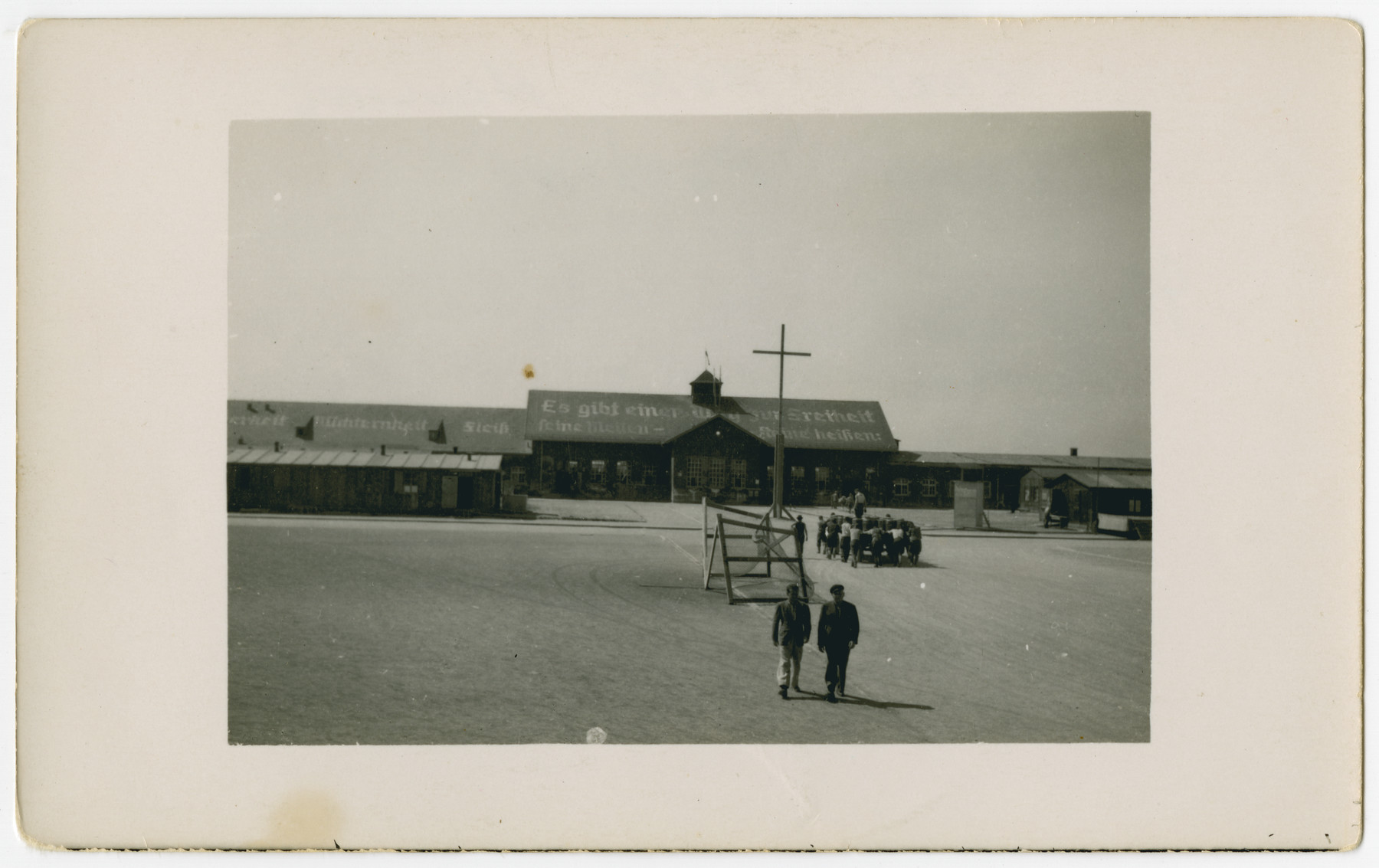 Two survivors walk through the central courtyard of the Dachau concentration camp while a larger group pushes a wagon in the background.

The original caption reads: "Cart for hauling dead prisoners."