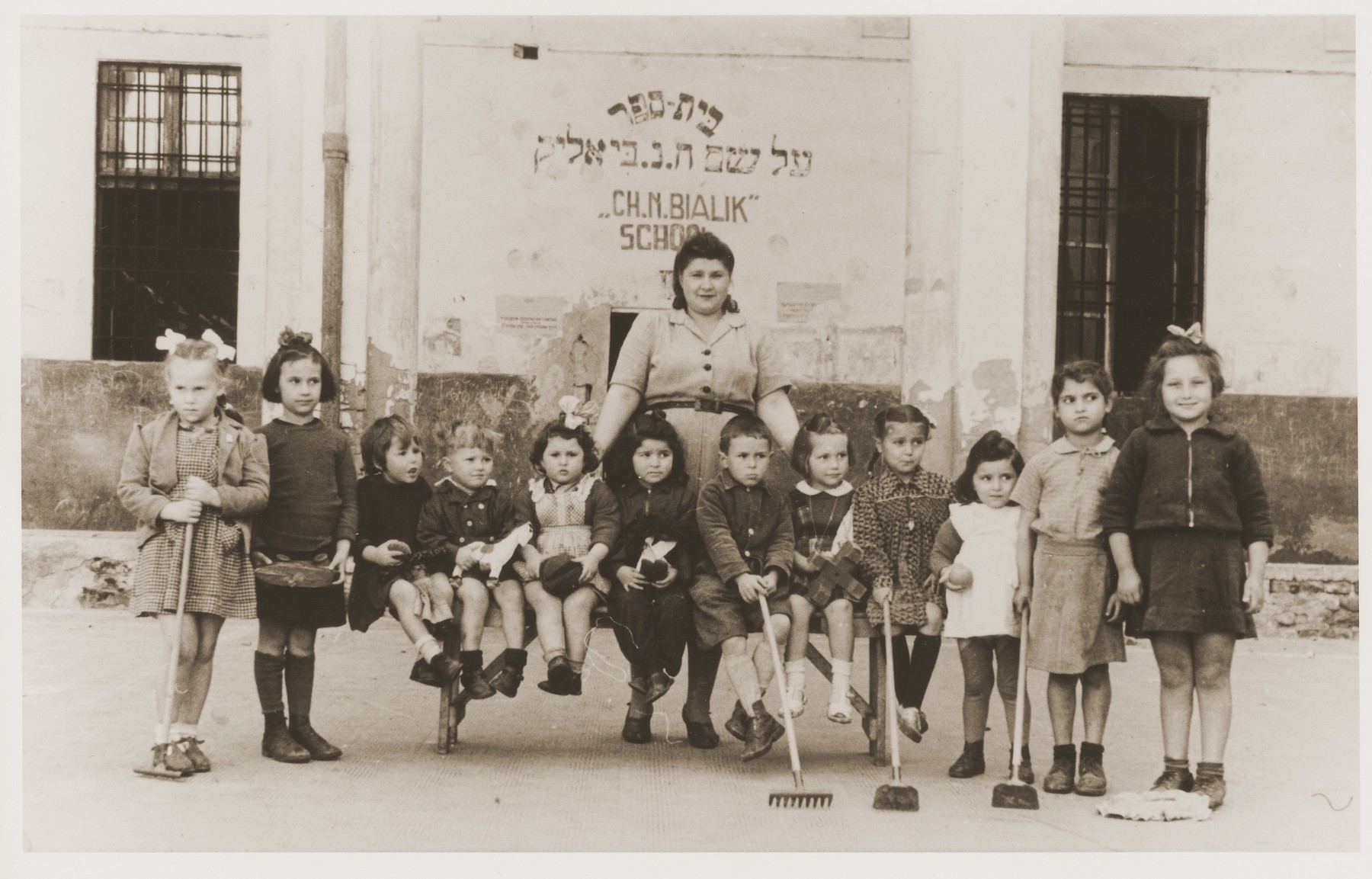 Jewish DP children at the Cremona displaced persons camp pose outside the Chaim Nachman Bialik school holding rakes and hoes.

Among those pictured is Masha Leikach (far left).