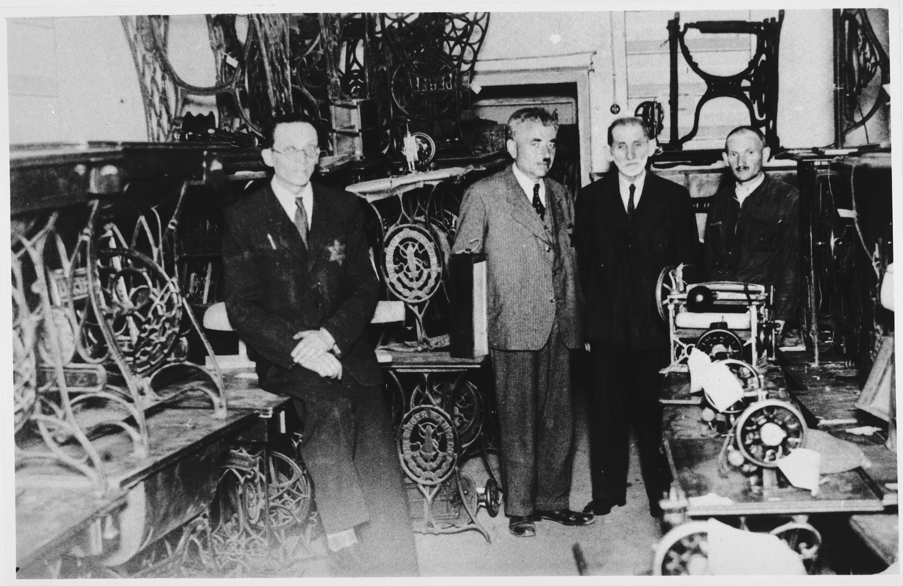 Four men wearing Jewish stars pose in a ghetto workshop filled with sewing machines.