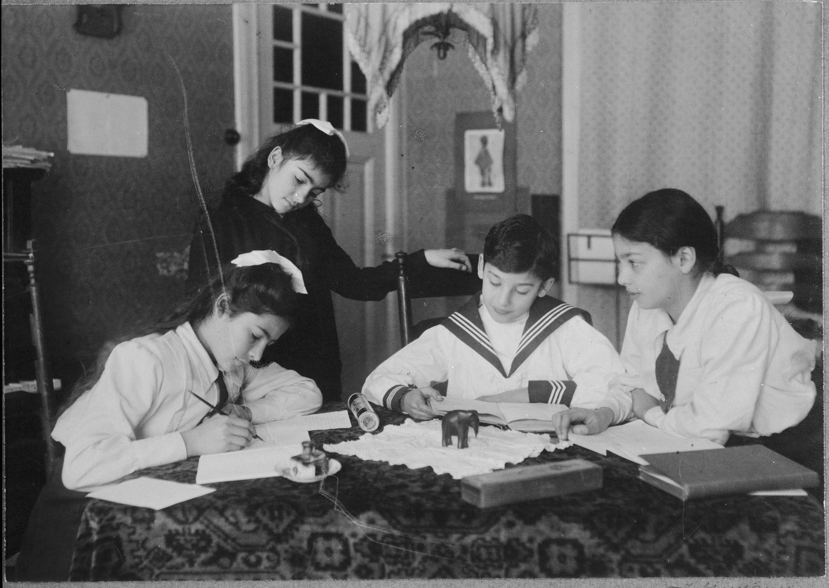 Four children gather around a table in their home.  

Pictured are Frieda Belinfante (standing), with her siblings Renee, Robert, and Dorothea (Dolly) (left to right).