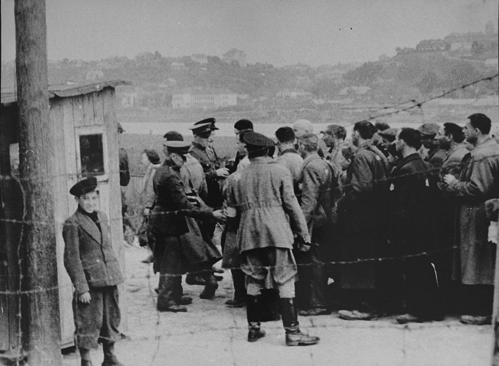 A group of Jewish men return to the ghetto after forced labor on the outside. 

Here, they line-up at the entrance to the ghetto to be searched by German and Lithuanian guards. A Jewish policeman stands with his back to the camera. A boy poses near the barbed-wire fence.  The Jewish men had to remove their hats in the presence of a German; the bald man in the center is Yankel Meishe Velikoludas.