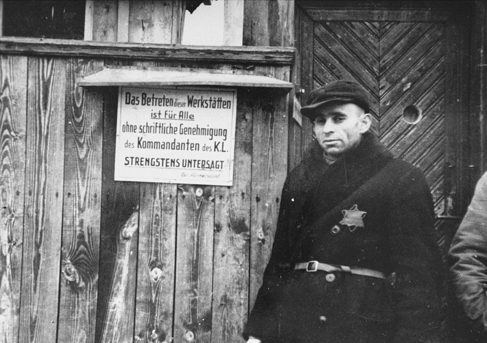 A Jewish man standing at an entrance to a workshop in the ghetto. The sign (in German) reads, "Entrance to this workshop is forbidden to everyone without a written permission of the camp Kommandant."