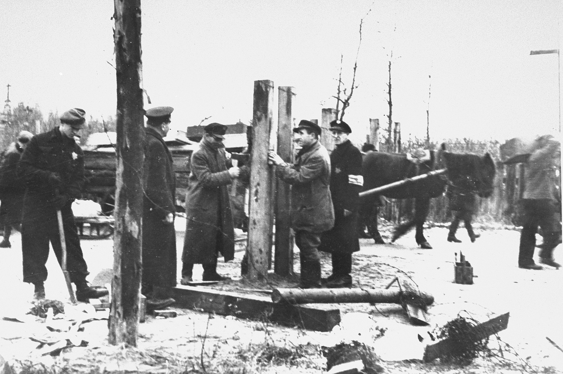 Jews repairing or moving the ghetto fence after a reduction of the ghetto boundaries.  Pictured at the left is Abe Malnik.  Second from the right is Yankel Kaplan.

On July 24, 1941, three weeks before all Jews had to move into the ghetto, the Kovno city government ordered that the Jews erect a fence around the ghetto.  A new order stipulated that work was to begin on July 30.  Fifty men were to report to work at 8:00 in the morning for the purpose of erecting the fence.  Posts were placed one meter apart and barbed wire was strung every twenty centimeters.  Signs were posted warning that the area  within three meters of the fence was declared a "death zone"; anyone caught in that zone would be summarily shot.  Many Jews were shot for approaching the fence.  However, when guards were not looking, Jews and Lithuanians traded through the fence, and some Jews, using wire cutters, managed to cut the fence and escape from the ghetto.  The ghetto fire department was responsible for mainaining and repairing the fence.