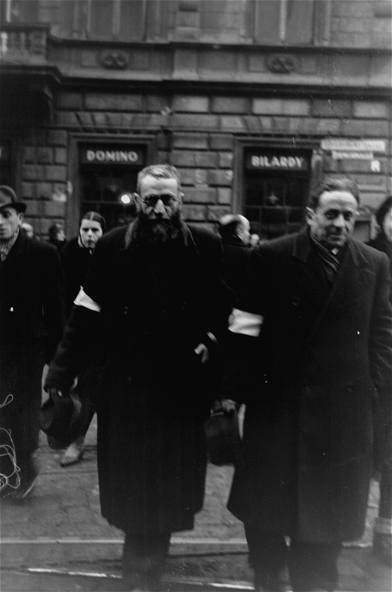 Jews crossing Chlodna Street in the Warsaw ghetto doff their hats to the photographer, in accordance with the German order requiring Jews to remove their hats in the presence of German personnel.