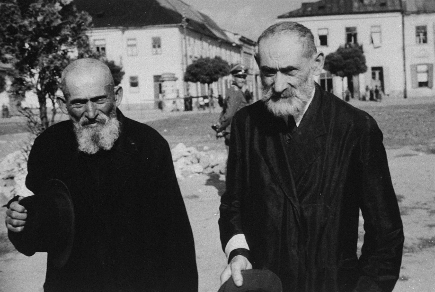 Two religious Jews wearing armbands are forced to remove their hats on a street in an unidentified ghetto.  

An SS officer is visible in the background.

The location of this photograph has been tentatively identified as Rynek Radom, Masovian Voivodeship, Poland.