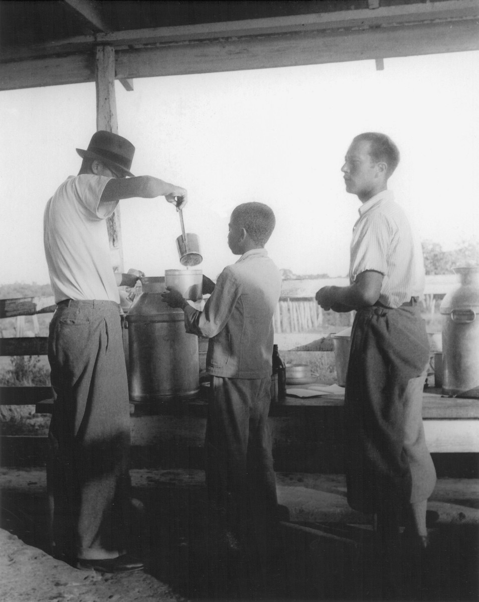 A Jewish refugee living in the Sosua settlement distributes milk to a Dominican youth in El Batey.  

Among those pictured are Alex Rohr (left) and Martin Katz (right).