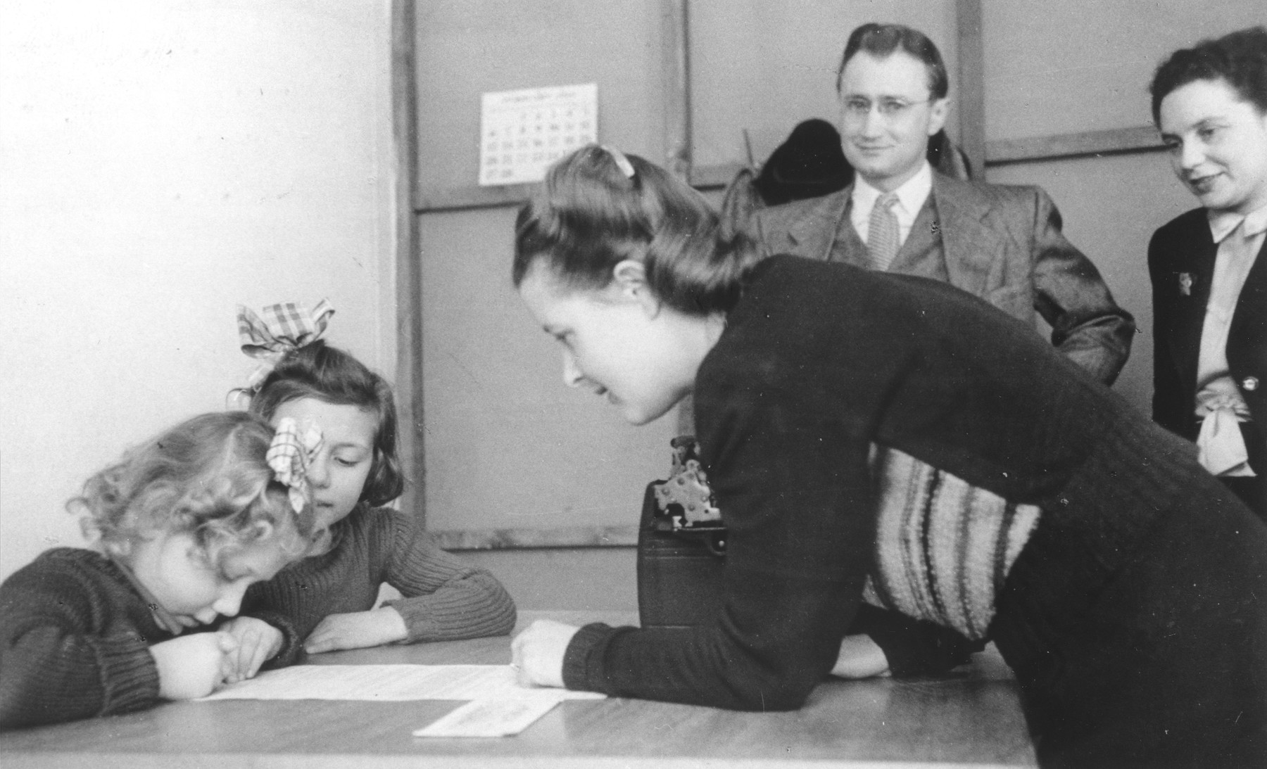 Syma Minc Klok, director of the Bad Schallerbach DP children's home, watches while seven-year-old Liesl signs her name on an American visa application.

Behind them stands Mr. Fischer, the American consul.