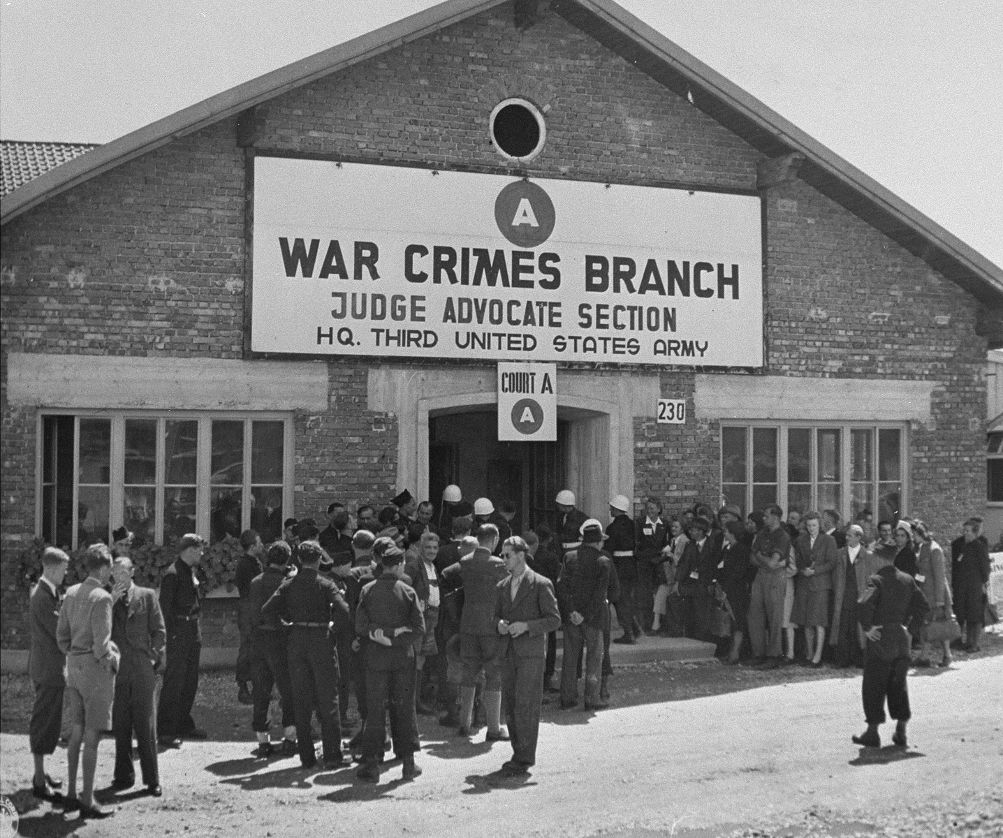 Spectators and U.S. Army military police wait outside of the building where 74 SS men will be sentenced for their roles in perpetrating the Malmedy atrocity.