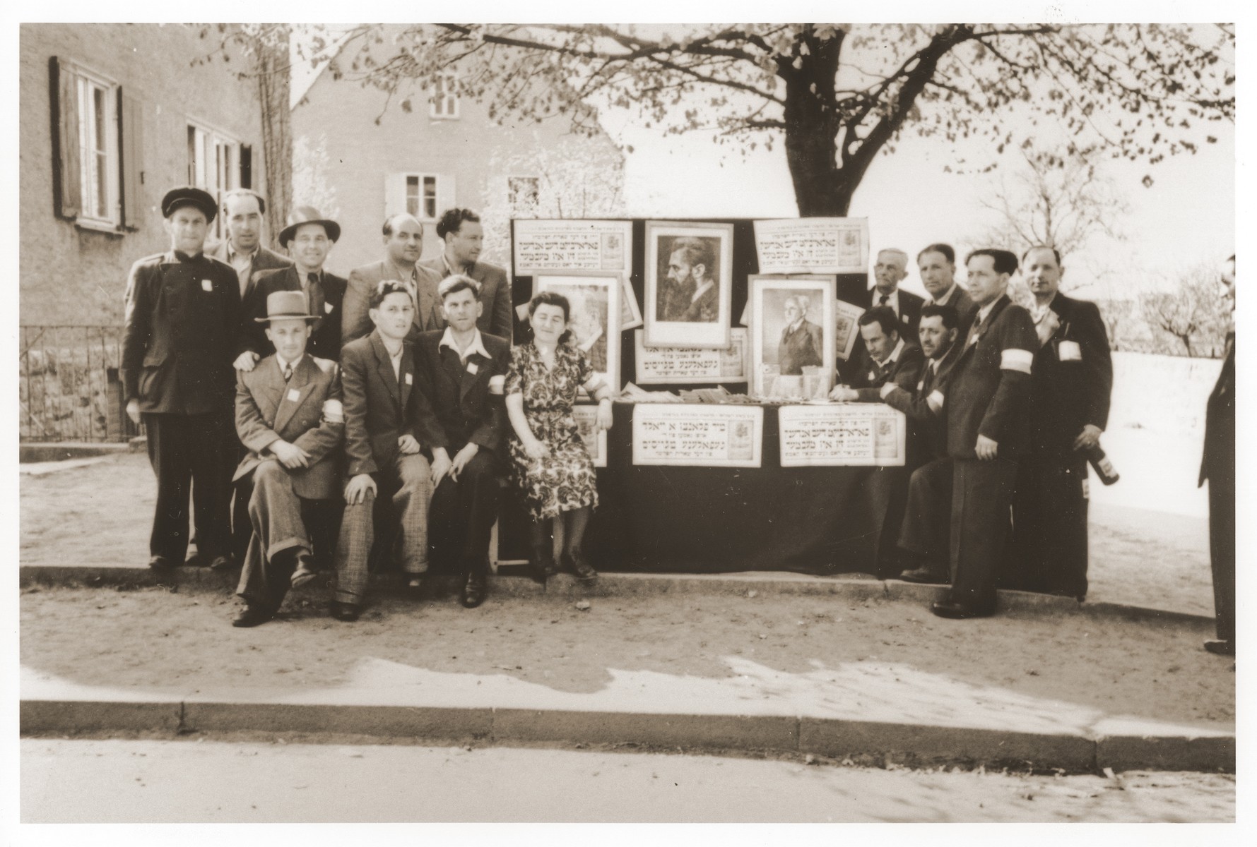 Members of the Hashomer Hatzair Zionist youth movement pose alongside an outdoor display in the Lechfeld displaced persons camp.

Gerzon Trzcina was born in Krasnosielc, Poland in 1922.  He was the son of Dina Kassel and Benam Yitzhak, who was a businessman in the leather trade.  Gerzon had one older brother, Jacob, and two sisters, Faiga (b. 1918) and Perl (b. 1920).  When the war broke out, Gerzon was working as a photographer's assistant (among other jobs) in Warsaw.  Following the German invasion of September, 1939, Gerzon was slightly wounded by shrapnel.  Soon after, a drunken German soldier, forcibly cut Gerzon's hair.  When the soldier made him stoop down to pick up the cuttings, Gerzon grabbed the German's gun and ran away.  After a short time in hiding, Gerzon returned to his hometown, where he learned that eighty Jews had been murdered in the synagogue during the first week of the war in one of the first large-scale atrocities against Polish Jews.  Among those killed was Gerzon's eighty-two-year-old grandfather, Haskel Kassel.  Gerzon moved on to Makow Mazowiecki, where he was forced to clean the stables the Germans had set up in the town's synagogue.  In the late fall, Gerzon escaped with his family to Bialystok, where he worked in a tannery until the family was deported to the Soviet interior in June, 1940.  Along with a group of Polish and Jewish refugees, the Trzcinas were sent to the Kiltovo labor camp in northern Russia.  Harsh conditions and poor nutrition led to Gerzon's father's death in the camp.  The family remained in Kiltovo until the German invasion of the Soviet Union in June of 1941.  Soon after, the refugees were allowed to leave the camp, and the Trzcinas moved to nearby Syktyvkar (capital of the Komi republic), where Gerzon worked in a construction company and later in a tannery.  In Syktyvkar, Gerzon was reunited with Gittel Blankitner, a Jewish refugee from Mlawa, whom he had met earlier in his travels in the Soviet Union.  (Gittel's father perished in Aushwitz, and her mother and siblings were killed in Treblinka.) In November, 1944, Gerzon and Gittel were married. The couple left the Soviet Union with their new baby in 1946.  Returning for a brief time to Poland, the family soon joined the wave of survivors streaming toward the Western zones of occupation.  After their arrival in Germany, the Trzcinas stayed in several DP camps including Vilseck, Wurzburg and Lechfeld, where Gerzon worked as an UNRRA photographer.  In March 1951, the family immigrated to the United States.