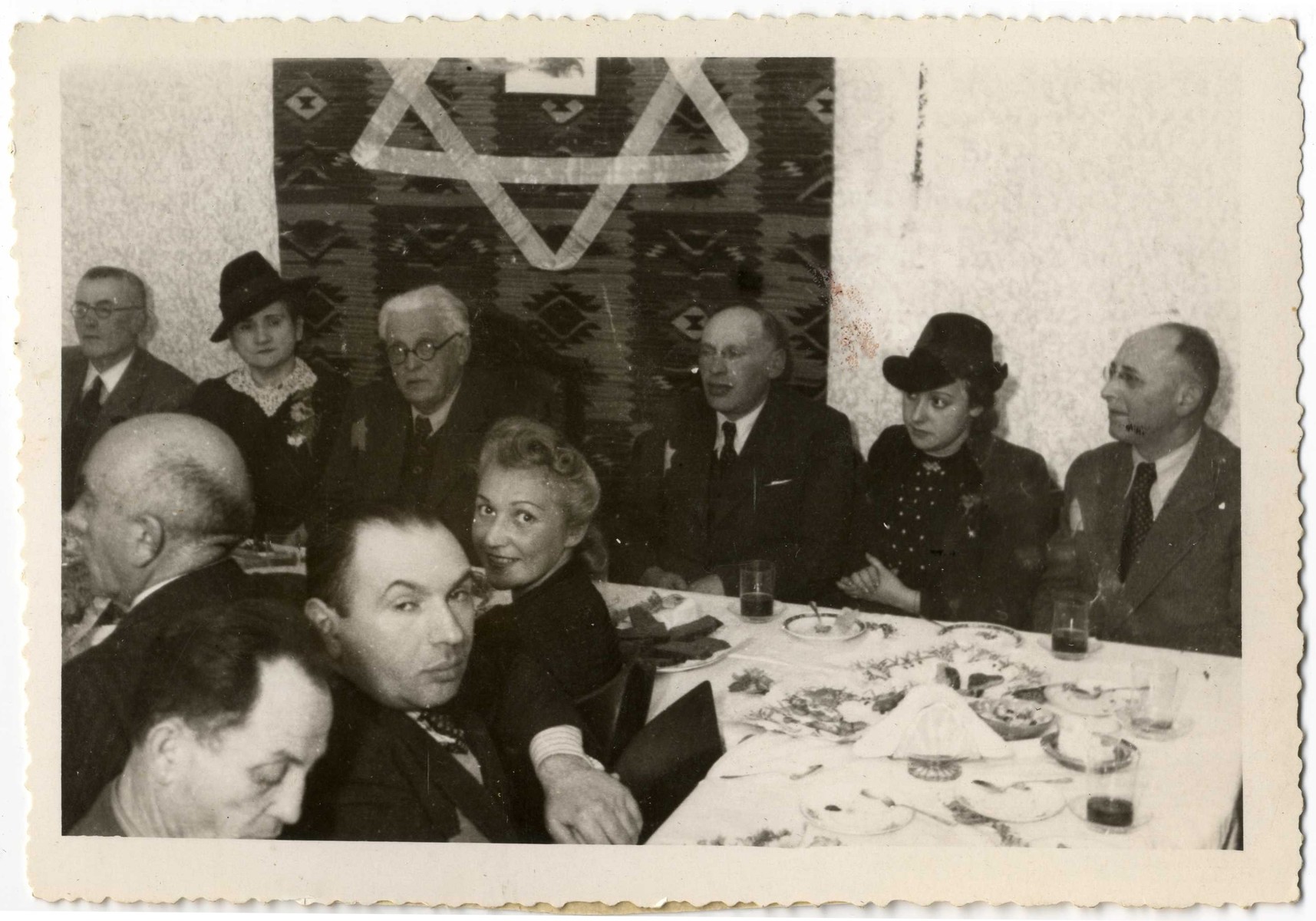 Ghetto officials gather for a festive meal in front of a wall hanging with a large Jewish star.

Seated in the center of the table is Chairman, Mordechai Chaim Rumkowski.  To his left is his sister-in-law Helena Rumkowski.  To his right are Dawid Warszawski and Dora Fuks.