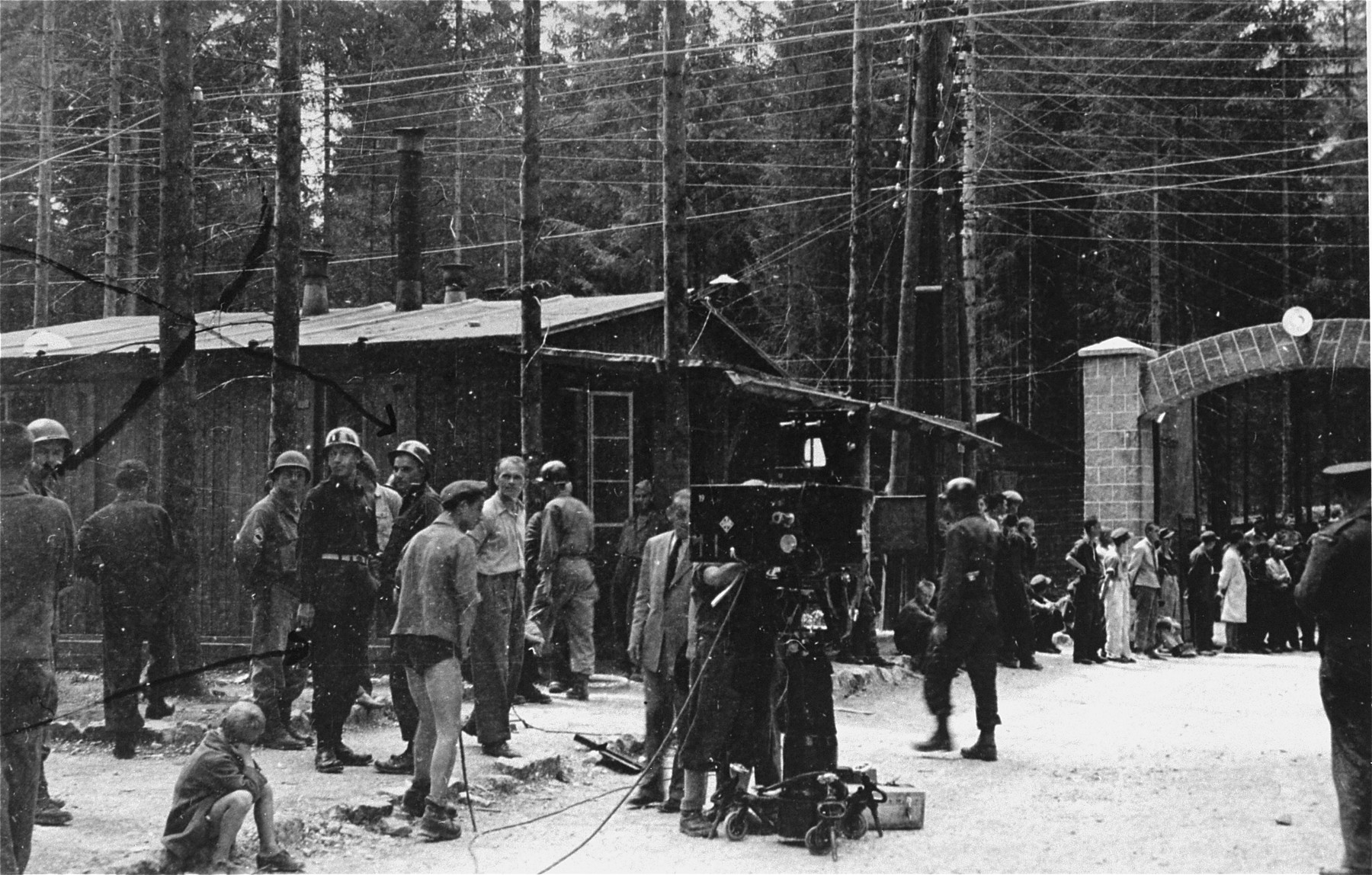 Survivors and American soldiers stand at the entrance to the Ebensee concentration camp and watch as a film crew sets up its equipment.